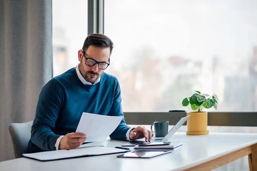 Bearded man checking piece of paper and laptop at desk